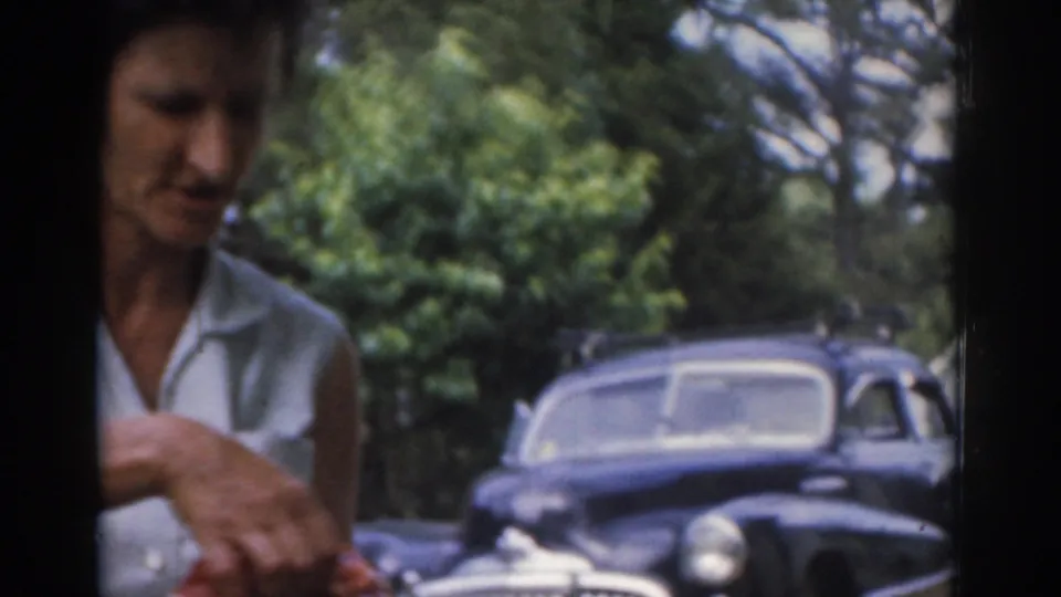 Close home-movie view of a woman with food in hand, with a vintage car and leafy outdoor setting behind her in New Jersey.