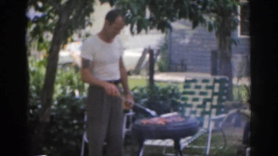 A man tends a small backyard charcoal grill near a green and white lawn chair in 1963 New Jersey home-movie footage.