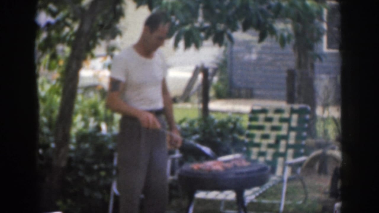 1963 New Jersey home-movie frame showing a man grilling meat beside a woven lawn chair in a leafy backyard.