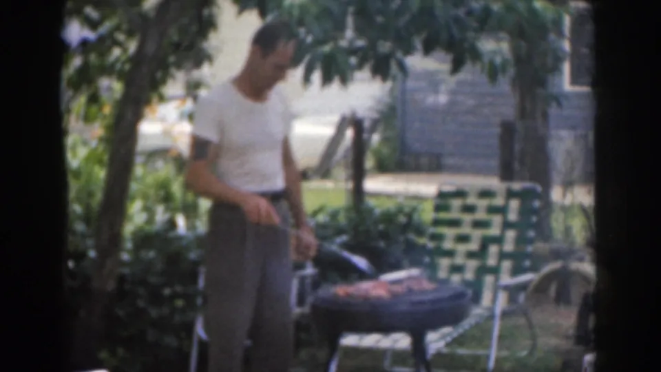 1963 New Jersey home-movie frame showing a man grilling meat beside a woven lawn chair in a leafy backyard.