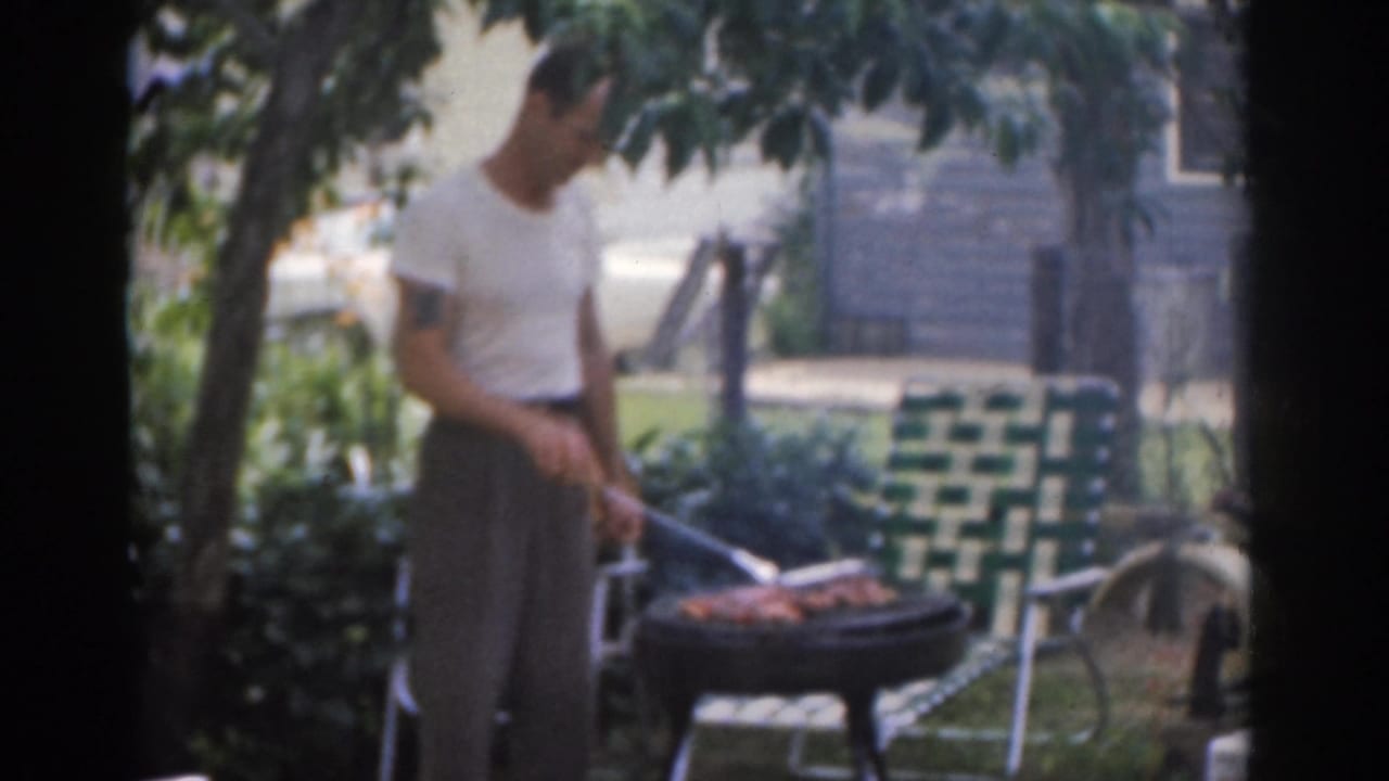 Man stands over a backyard grill with meat cooking near a lawn chair.