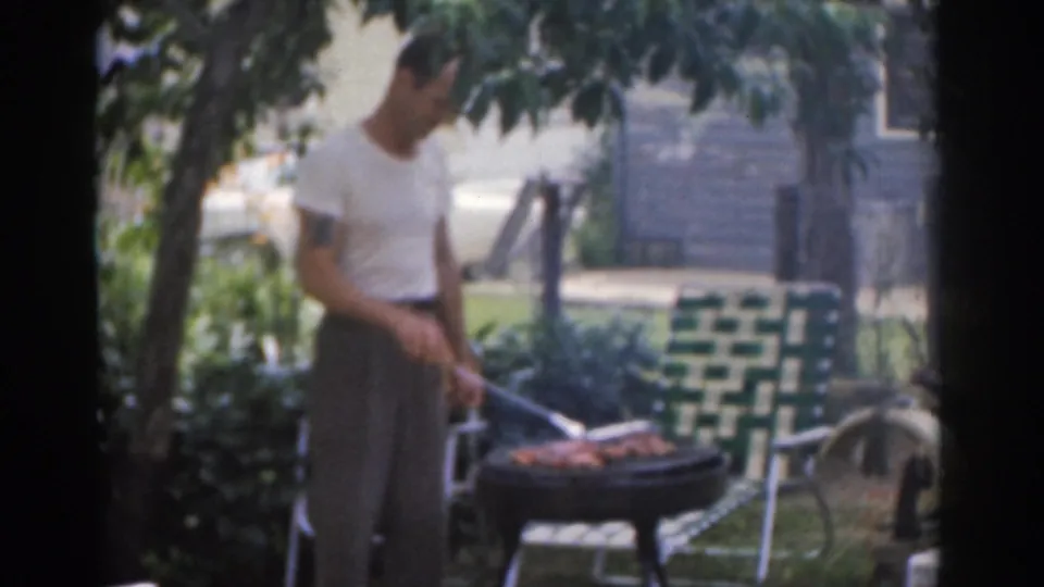 The man continues grilling chicken and beef in a leafy New Jersey backyard, with a green-and-white lawn chair nearby.