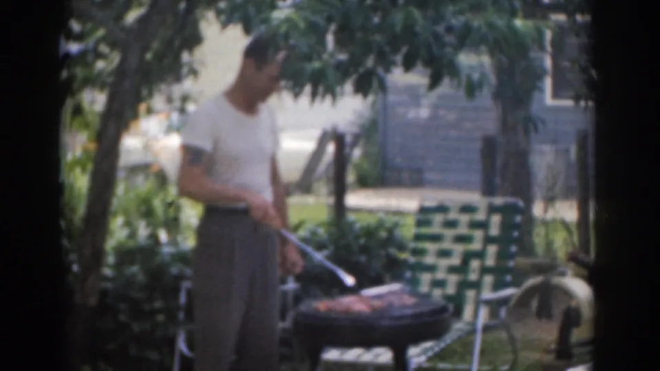 A 1963 8mm frame shows the man pausing with a grilling utensil beside the barbecue, with lawn furniture and foliage behind him.