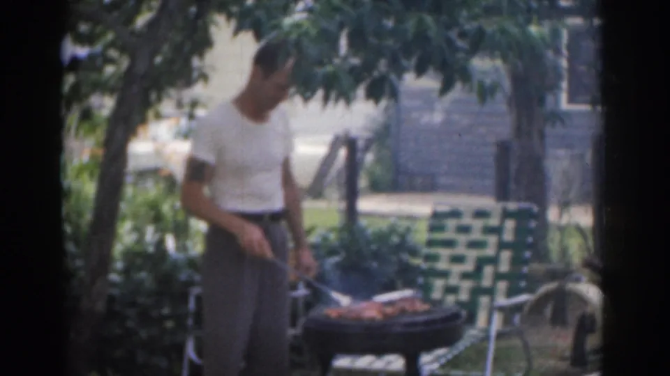Final frame of the short 1963 New Jersey barbecue scene, with the man looking down at the grill in a leafy residential yard.