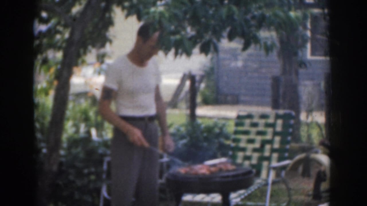1963 New Jersey 8mm frame of a man in a white shirt grilling in a shaded backyard with smoke and a lawn chair nearby.