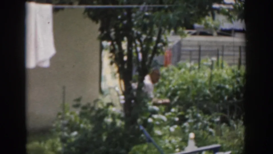 1963 New Jersey 8mm frame showing a shaded garden, hanging towel, fencing, and a person partly obscured among the plants.