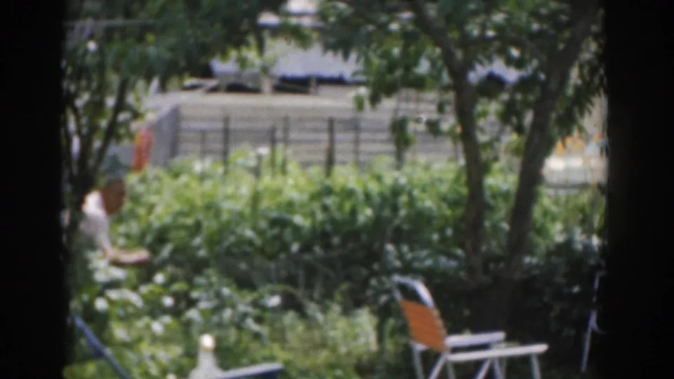 1963 New Jersey home-movie frame showing a leafy backyard with folding chairs, garden foliage, and a person partly visible near the fence.