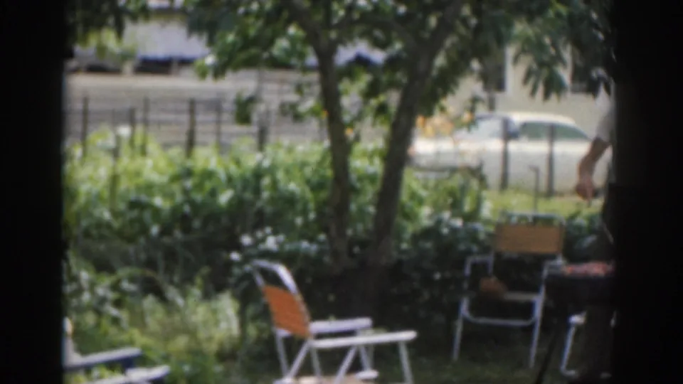 1963 New Jersey home-movie frame showing a shaded barbecue area with folding chairs, trees, grill equipment, and an adult partly in view.