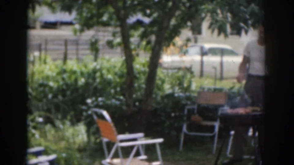 1963 New Jersey backyard barbecue frame with folding chairs, a grill setup, a seated adult, trees, and a white car beyond the fence.