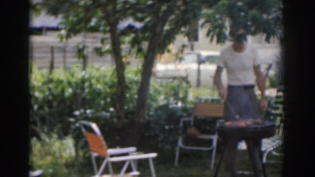 Man standing by a charcoal grill in a leafy backyard with lawn chairs nearby.