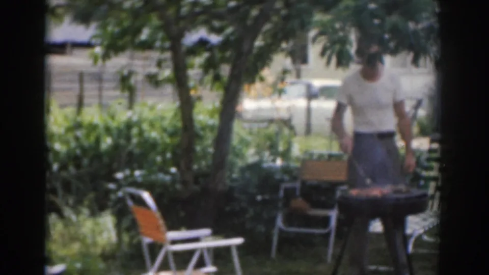 1963 New Jersey home-movie frame showing a man by a charcoal grill under trees in a backyard with folding chairs.