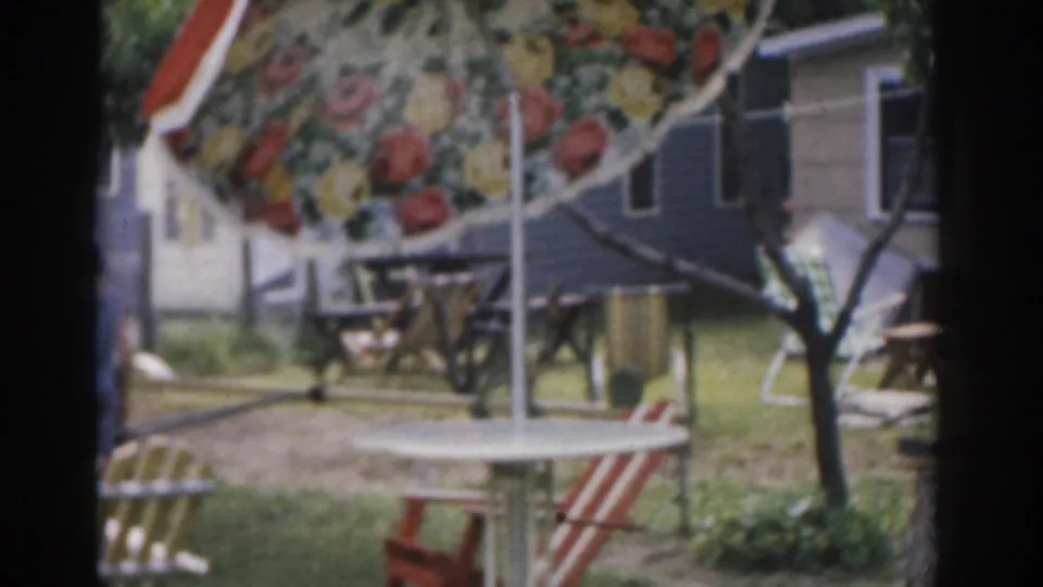 1963 New Jersey backyard view with a patterned floral umbrella, picnic table, folding chairs, and house exterior.