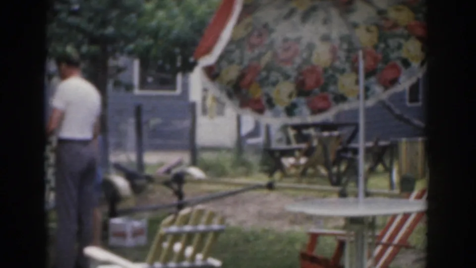 1963 New Jersey home-movie frame showing a man from behind near a charcoal grill in a backyard picnic area.