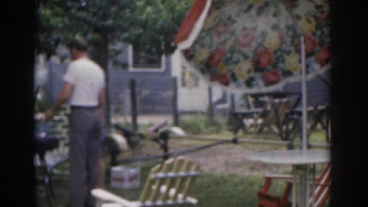 Man in a white shirt stands near a backyard grill beneath a floral patio umbrella.