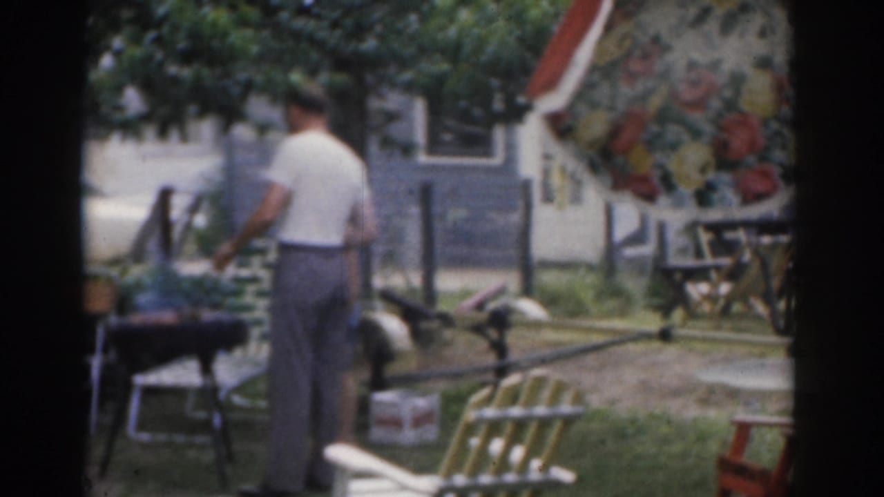 Man stands by a grill in a yard with a floral umbrella and house in the background.