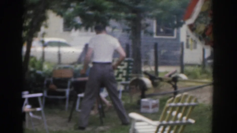 1963 New Jersey frame showing a man moving through a backyard barbecue area with chairs, floral umbrella, white fence, and a car in the background.