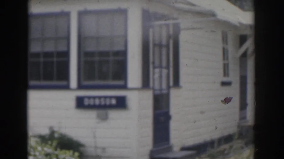 1963 New Jersey home-movie frame focused on the exterior of a light-colored residential house with windows, a screen door, and a small sign or plaque.