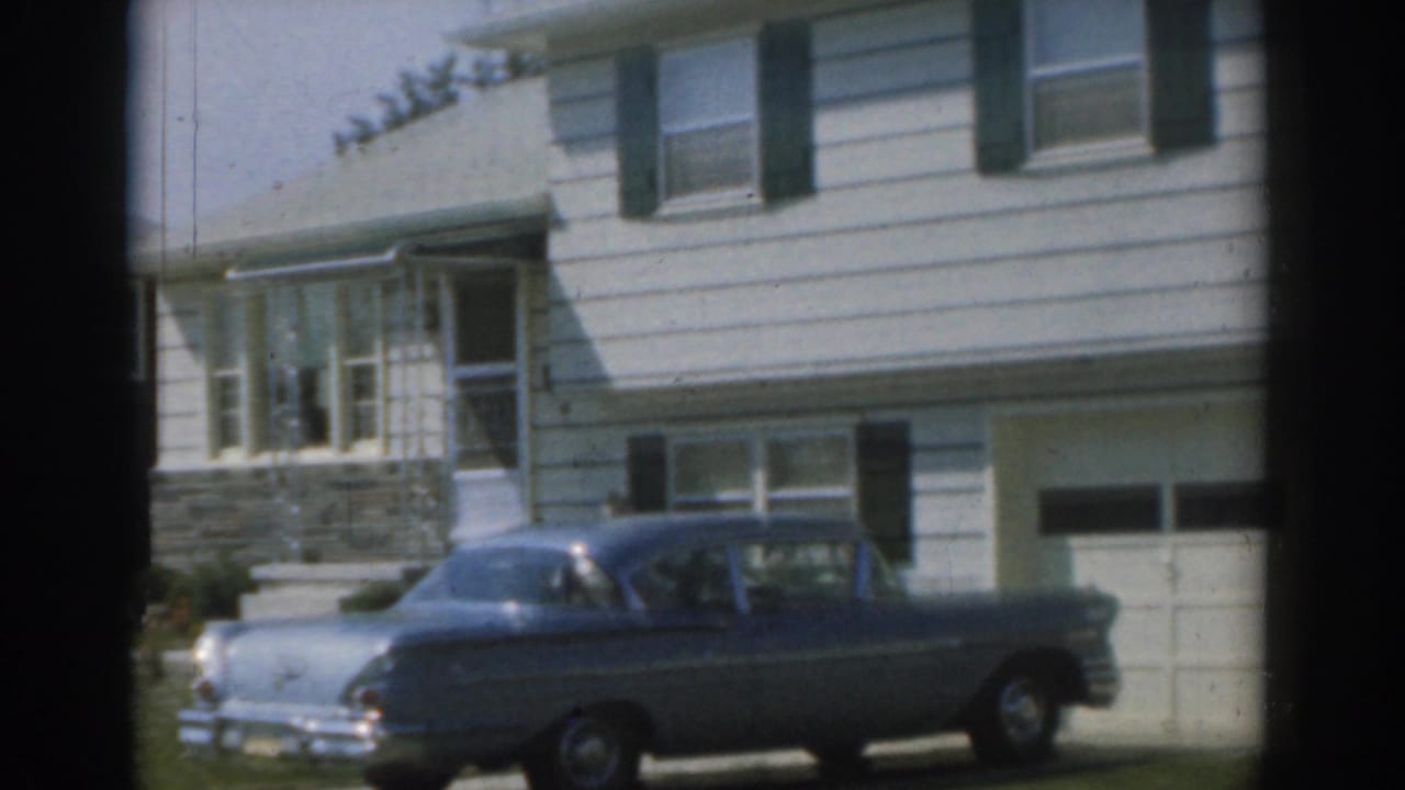 Blue sedan parked in front of a white suburban house with green shutters.