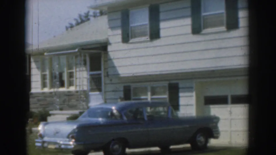 1963 New Jersey home-movie frame showing a blue sedan beside a white house with green shutters, windows, stone trim, and lawn.