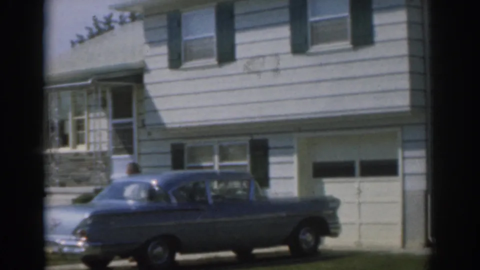 1963 New Jersey frame with a man partly visible near a parked blue sedan in front of a white house and garage.