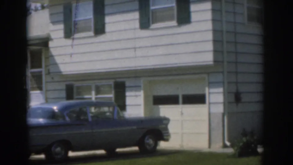 Home-movie frame from 1963 New Jersey showing the blue sedan lower in frame beside a white house, garage door, shutters, and lawn.