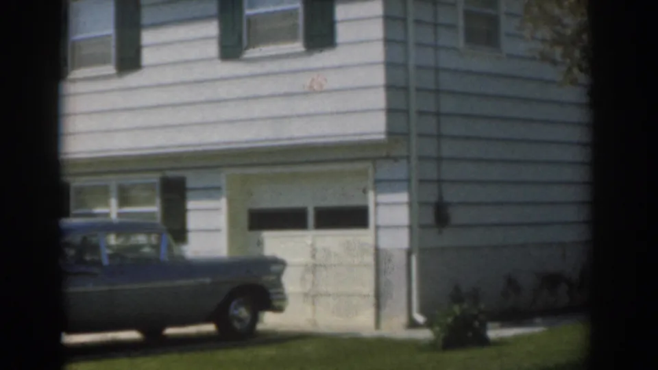 1963 New Jersey home-movie frame with the sedan partly cropped at left, a white house facade, garage door, windows, and lawn.