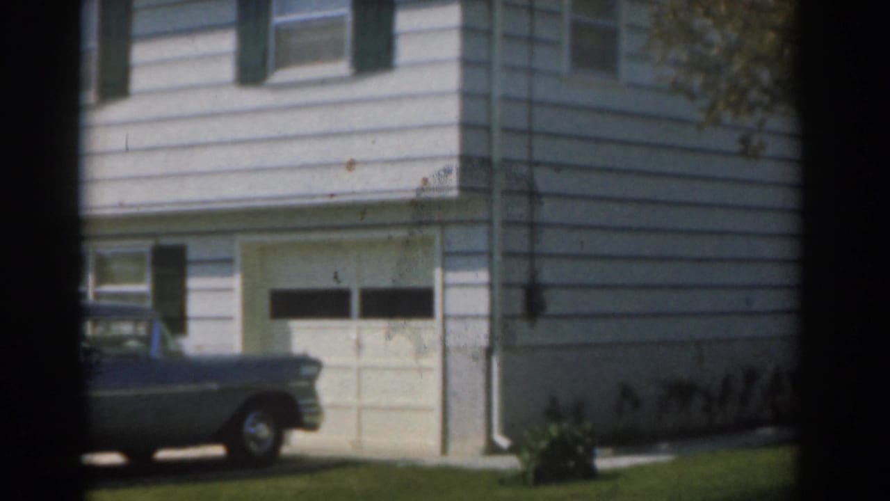 Grey sedan parked beside a white-sided house with a garage door and green shutters.