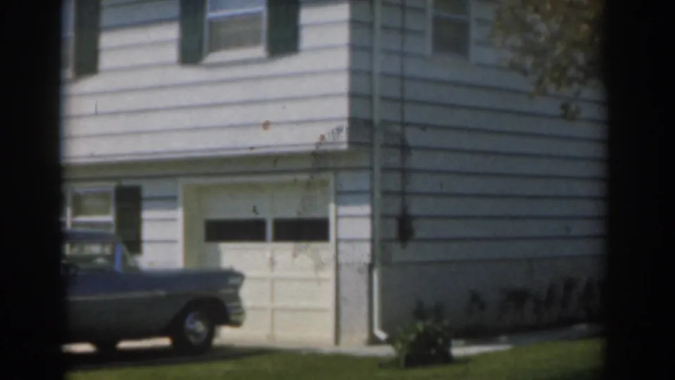 1963 New Jersey home-movie frame showing a grey sedan near a white suburban house, garage door, green shutters, and lawn.