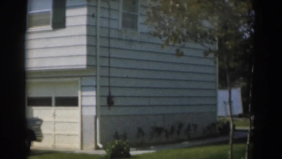 1963 New Jersey frame with the camera panning across a white suburban house, garage door, lawn, and partial grey car.