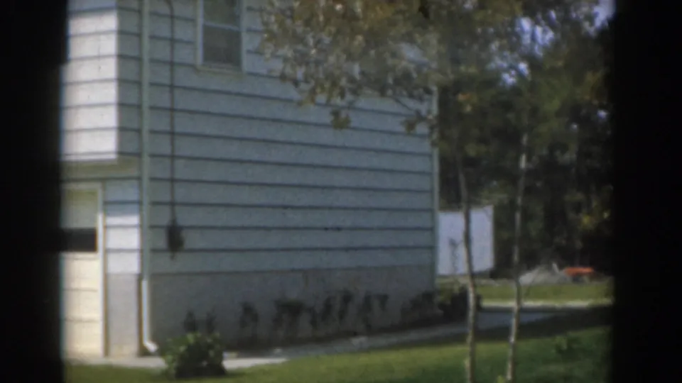 1963 New Jersey home-movie frame showing the side of a white suburban house with lawn, shrubs, young trees, and a clothesline area.
