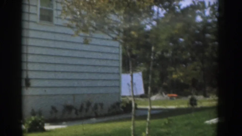 1963 New Jersey frame showing a sunny suburban side yard with white house siding, young trees, lawn, and a clothesline in back.
