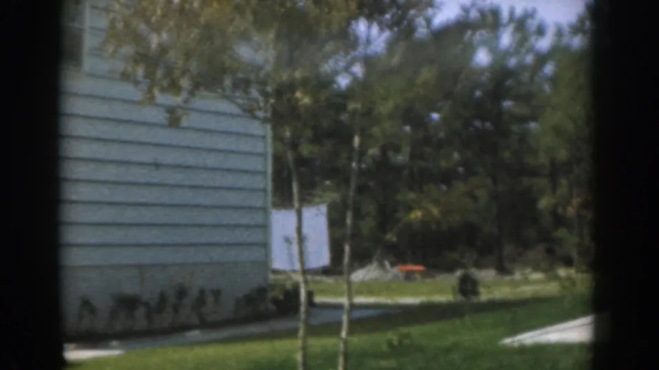 1963 New Jersey 8mm frame showing a street-side residential yard with white siding, lawn, trees, and a distant parked car.