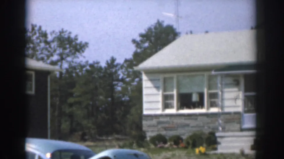 1963 New Jersey residential exterior with a white house, front window, porch area, shrubs, mature trees, and parked cars at street level.