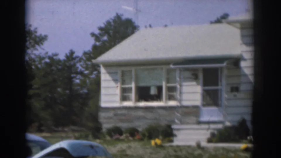 1963 New Jersey 8mm view of a white two-story house front, showing porch steps, a large window, low shrubs, lawn, and a car roof.