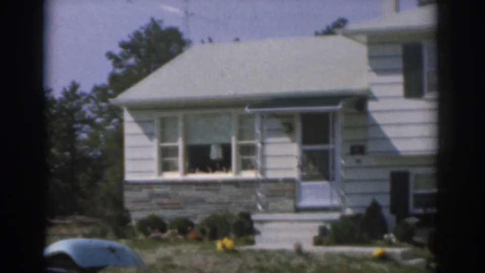 1963 New Jersey frame of a white house exterior with covered porch, front steps, picture window, shrubs, lawn, and part of a parked car.