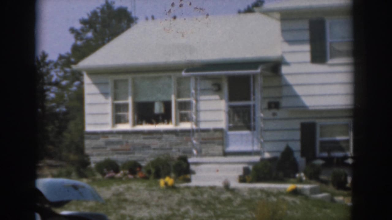 White suburban house with front lawn, awning, shrubs, and partial parked car.