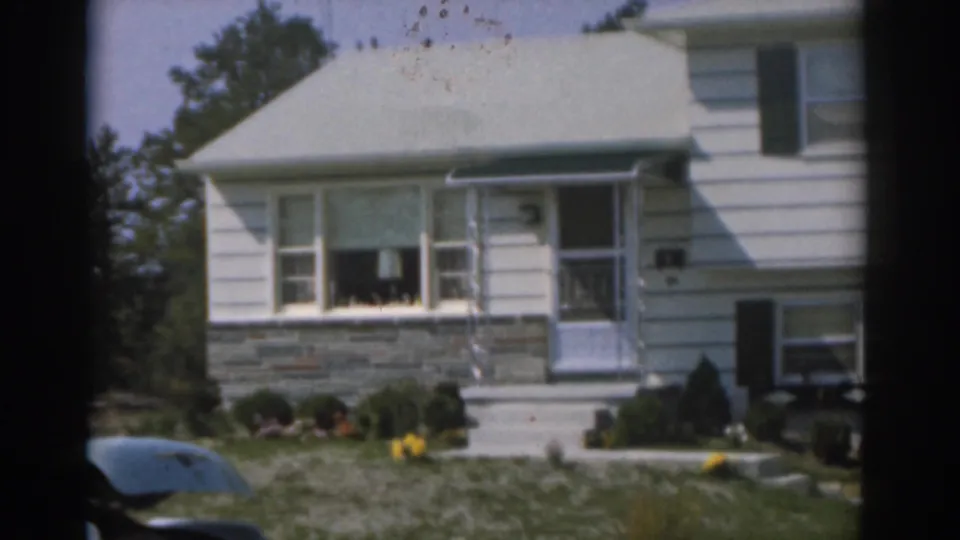 1963 New Jersey 8mm view of a white suburban house with stone facade, green shutters, front lawn, and a car partly visible at left.