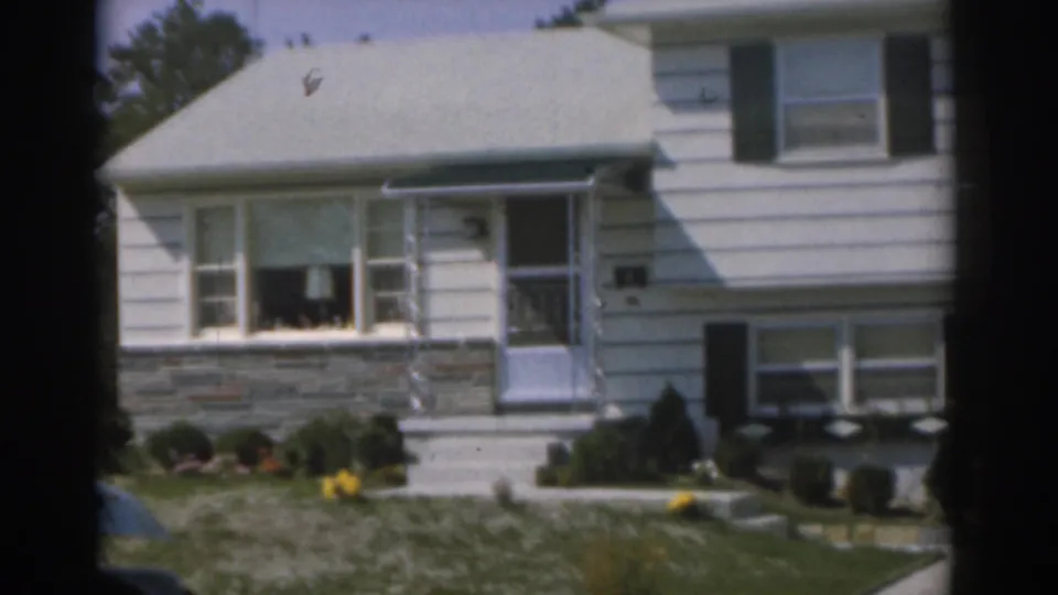1963 New Jersey home-movie frame showing the front of a white suburban house, stone trim, shaded doorway, lawn, and garden plantings.