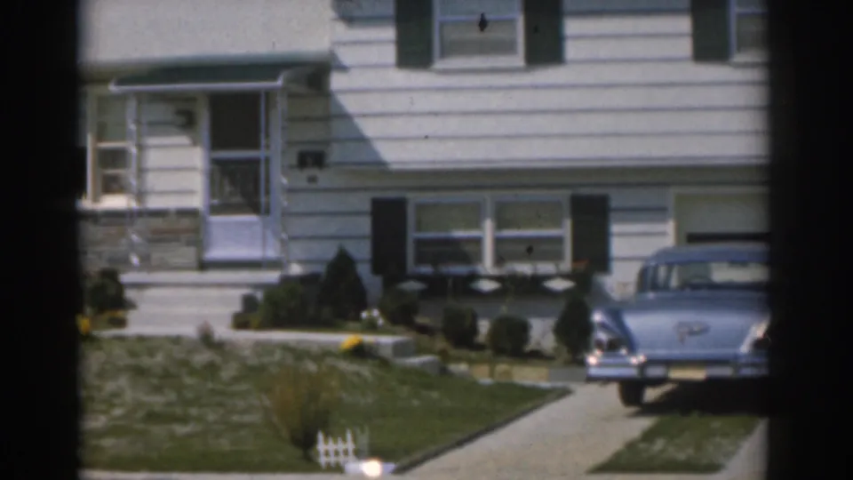1963 New Jersey frame showing the suburban house exterior, driveway, parked 1960s car, lawn, shrubs, and front entrance.