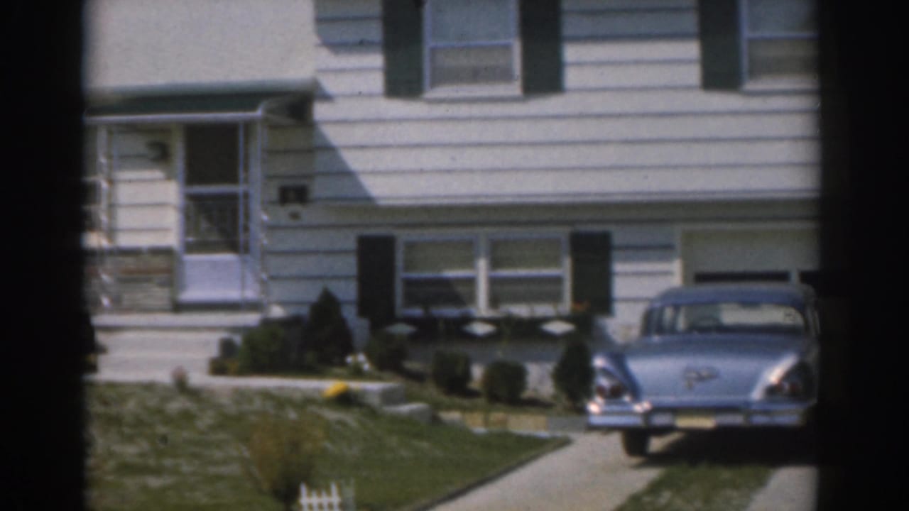Blue sedan parked in a driveway outside a white suburban house with dark vertical frame edges.