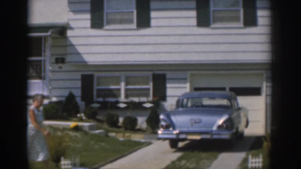 A woman in a light dress crosses a New Jersey suburban lawn near a blue 1960s sedan and white-sided house.