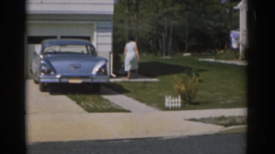 The woman continues across the suburban lawn, seen beside the driveway, garage, small fence edging, and parked blue sedan.