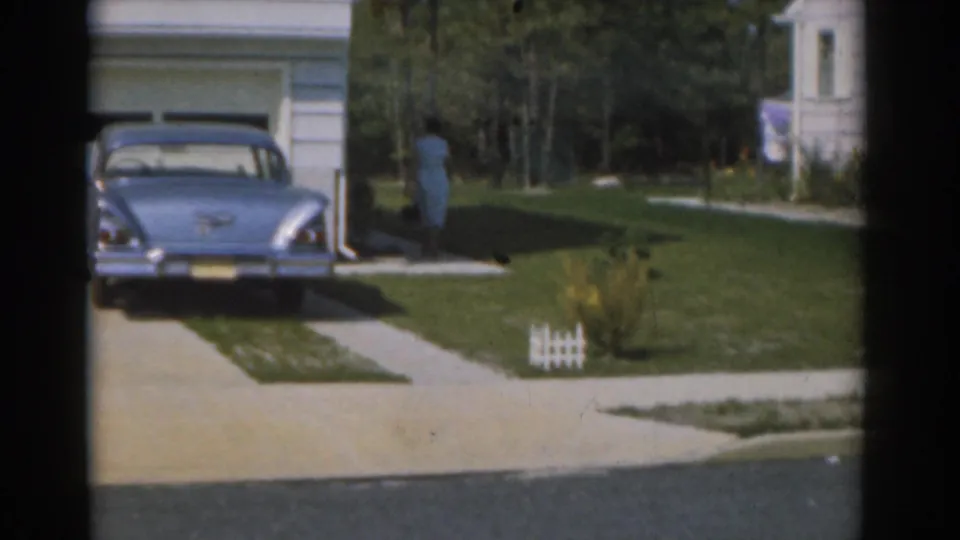 Near the end of the shot, the woman is farther right on the lawn while the blue sedan remains parked by the garage.