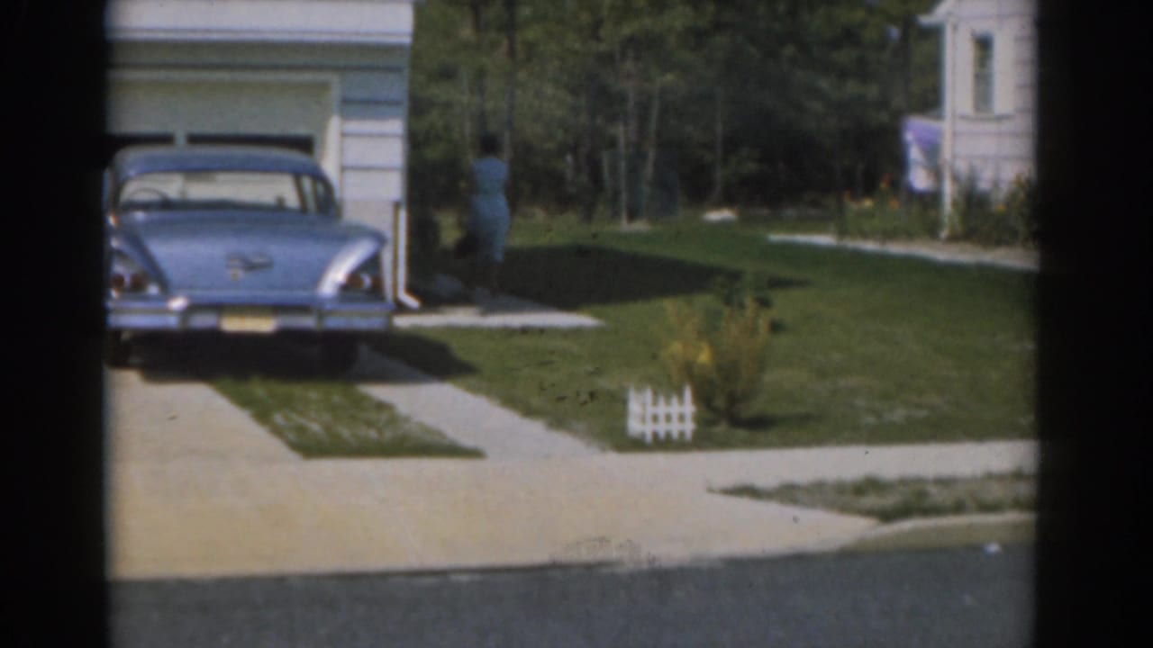 Blue vintage sedan parked in a driveway beside a garage, with a woman near the house.
