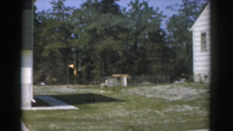 1963 New Jersey home-movie frame showing a suburban side yard with white house siding, trees, lawn, and driveway pavement.
