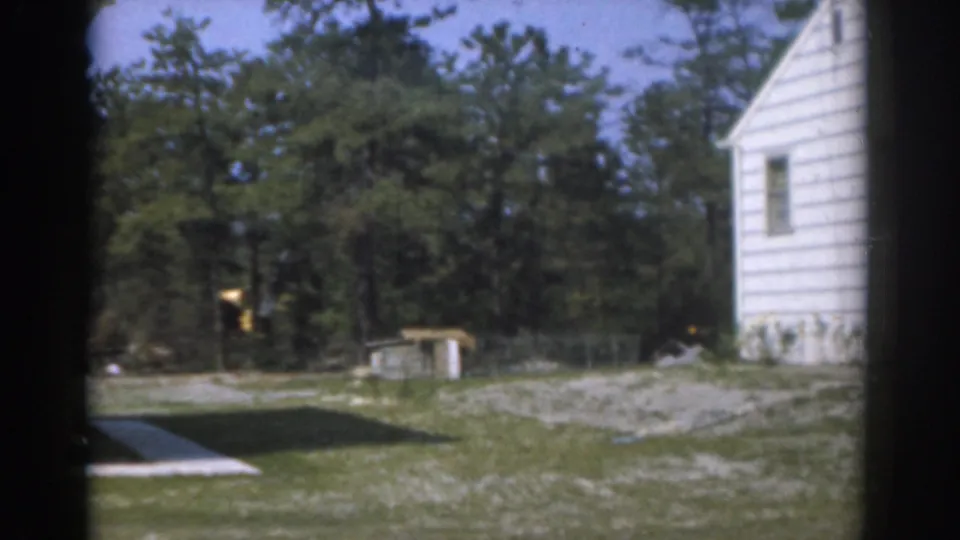 1963 New Jersey home-movie frame of a quiet residential side yard with white siding, trees, grass, and a small yard structure.