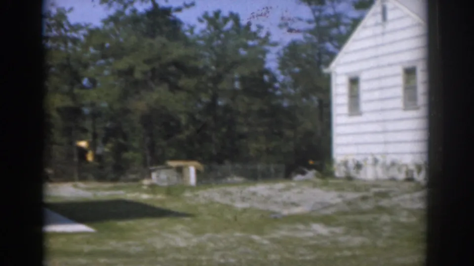 1963 New Jersey home-movie frame showing a suburban yard, white house wall, trees, and driveway edge in soft 8mm color.