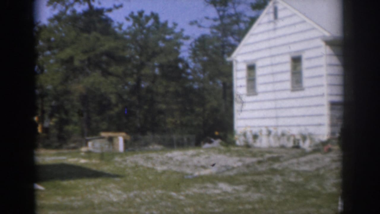 White clapboard house partly framed by dark film gate edges, with lawn, gravel, shed, and trees.