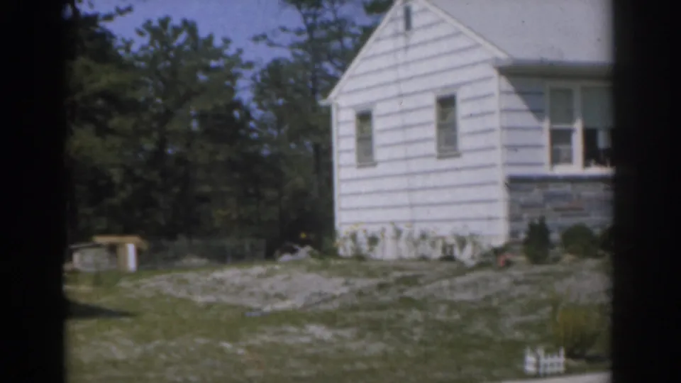 1963 New Jersey frame of a white suburban house with stone foundation, surrounding trees, lawn, gravel, and a small shed.