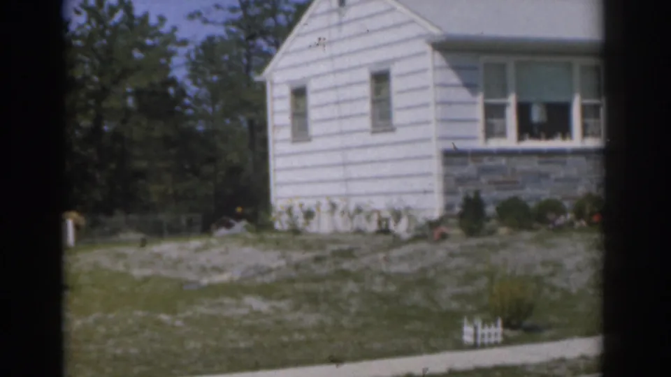 1963 New Jersey home-movie frame showing a white house, porch windows, stone base, sidewalk, lawn, and small white fence.